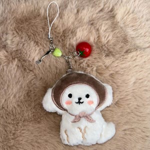 May include: A white plush dog charm wearing a brown hat and a pink bow. The charm has a silver chain with a fork charm, a green heart, and a red tomato charm. The background is a fluffy, light brown surface.