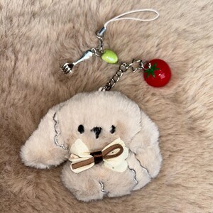 May include: A beige, dog-shaped plush charm with a cream and brown bow tie. It hangs from a silver chain with a tiny fork, green bead, and a red tomato charm. The background is a soft, brown, furry texture.