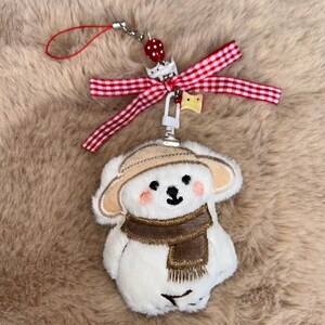 May include: A white plush bear charm with a brown scarf and hat, featuring rosy cheeks and a black nose. A red and white gingham bow and a strawberry bead add detail to the keychain.