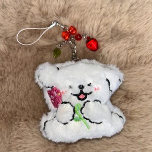 May include: A white plush bear keyring with black embroidered features, holding a green stem and a pink strawberry. The keyring has a silver chain with red bead accents and a red strawberry charm.