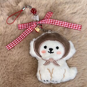 May include: A white plush dog charm wearing a brown hat and rosy cheeks. It's attached to a red and white gingham ribbon, a red string, and a metal clasp. A red strawberry bead and a small white dog charm are also attached.