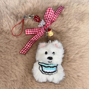 May include: A white plush bear keyring with a blue bib, black eyes, and pink ears. It has a red and white gingham ribbon, a strawberry charm, and a small bell charm. The keyring is attached to a red string.