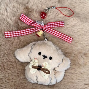 May include: A beige plush dog charm with black eyes and a bow. It features a red and white gingham ribbon and a strawberry charm. The charm is attached to a silver keychain and a red string.