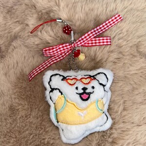 May include: A white plush bear charm wearing sunglasses and a yellow vest. It features a red and white gingham ribbon and a red wrist strap. A small chain and a strawberry charm are also attached.