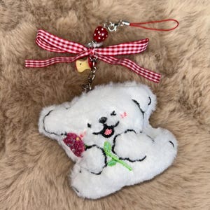 May include: A white plush bear keyring holding a pink flower. The bear has black embroidered features and a smiling face. It's attached to a red and white gingham ribbon and a red wrist strap. The background is a fluffy, brown texture.
