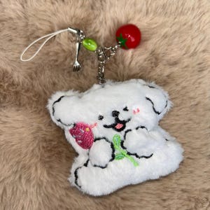 May include: A white plush dog charm with black outline details, holding a pink strawberry and green stem. The charm has a red tomato and green beads attached to a silver chain. A small silver fork charm and white loop are also attached.