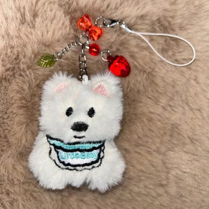 May include: A white plush bear charm with black eyes and nose, pink ears, and a blue bib with text. The charm is attached to a silver chain with red and orange beads, a green leaf, and a red strawberry charm.