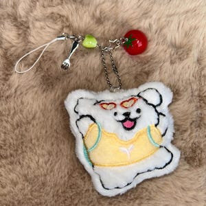 May include: A white plush bear keyring with a yellow vest and heart-shaped sunglasses. The keyring has a silver fork charm, a green bead, a red tomato charm, and a white cord.