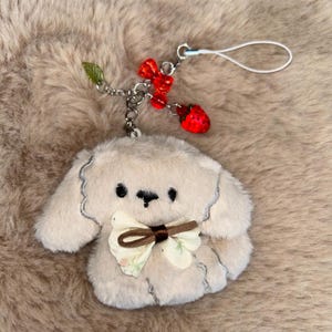 May include: A beige puppy plush keychain with a brown bow and floral details. It has a silver chain with red bead and strawberry charms, and a white loop for attachment.