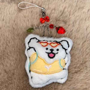 May include: A white plush bear-shaped keyring with black outlines, wearing heart-shaped sunglasses and a yellow top. The keyring has a red bow, strawberries, and a green leaf charm attached.