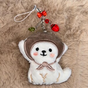 May include: A white plush bear charm wearing a brown hat and scarf. The charm features a silver chain with a red bow, red beads, a green leaf, and a red strawberry charm. The bear has pink cheeks and embroidered details.