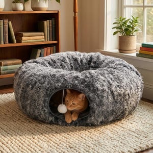 Puede incluir: Una cama para gatos gris y afelpada con forma redonda y una pequeña abertura. Un gato naranja está dentro de la cama, mirando hacia afuera. Un juguete de pompón blanco cuelga de la abertura. La cama está sobre una alfombra beige.