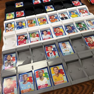 May include: A plastic sorting tray containing numerous rectangular soccer player trading cards. The cards feature portraits of athletes in various team jerseys, organized in rows within white and gray compartments. Each card displays a player's name.