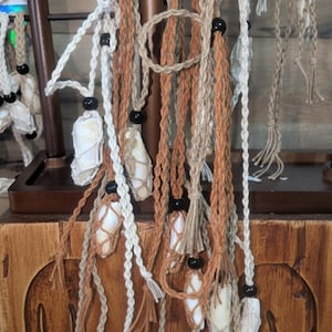 May include: A display of handmade necklaces featuring natural shells encased in macrame netting. The necklaces are suspended from a wooden stand. The macrame cords are in shades of white, tan, and brown, accented with black beads.