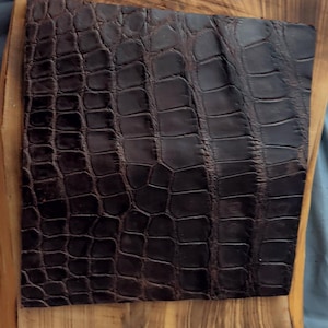 May include: A square piece of dark brown leather with a textured crocodile skin pattern. The leather is positioned on top of a light brown wooden board that features a natural grain pattern and an irregular, organic edge shape.