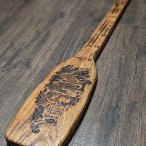 May include: A wooden paddle with a natural finish featuring a wood-burned design of a swamp scene, a crawfish, and the text "LAISSEZ LES BONS TEMPS ROULER". The handle includes depth markings in feet, starting at 2 ft and extending up to 3.5 ft.