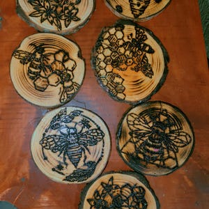 May include: A set of seven circular wooden coasters featuring intricate pyrography designs of bees, honeycombs, and floral patterns. Each coaster displays a unique wood-burned illustration on a light-colored wood slice with natural bark edges.