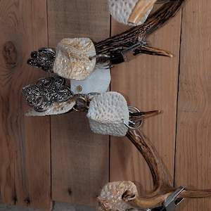 May include: A wall-mounted display featuring brown deer antlers attached to vertical wooden planks. Several white, textured, porous clay shapes with gold leaf accents are suspended from the antlers using silver metal key rings and black clips.