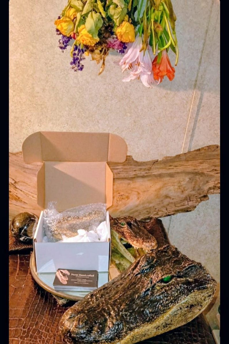 May include: A small, open cardboard shipping box containing bubble wrap and white tissue paper, accompanied by a rectangular business card labeled "Pura Handcrafted," positioned next to a realistic, textured alligator head sculpture with green eyes.