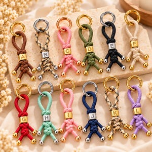 Può includere: Una collezione di dodici portachiavi in paracord a forma di piccole figure, ognuno dotato di un anello in metallo dorato e una perlina quadrata stampata con una lettera dalla A alla L. I cordini sono disponibili in vari colori e motivi.