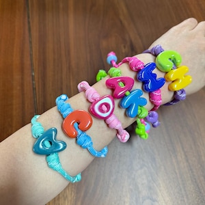 May include: A collection of colorful bracelets made from braided nylon cord, each featuring a single large, glossy, letter-shaped bead in various vibrant colors including teal, orange, pink, blue, green, and yellow, arranged in a stack.