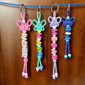 Op de afbeelding: Vier gepersonaliseerde sleutelhangers met de namen: "RACHEL", "MARY", "MEGAN" en "BRIA". Elke sleutelhanger heeft een decoratief knoopontwerp in roze, groen, lavendel en blauw, met bijpassende kralen en hartaccenten. De namen zijn gespeld met kleurrijke letterkralen.