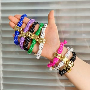Pode incluir: Uma coleção de pulseiras de corda coloridas com fechos dourados. As pulseiras apresentam uma variedade de cores, incluindo azul, roxo, preto, verde, branco, rosa e preto. Cada pulseira tem um fecho dourado com um design de letras.