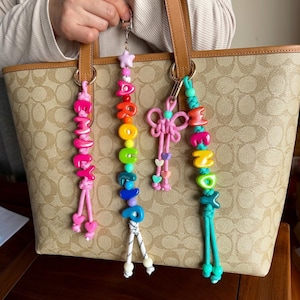 May include: A collection of colorful beaded keychains with names and decorative elements. The keychains feature a variety of colors, including pink, orange, yellow, green, blue, and turquoise. Each keychain has a unique design, with some spelling out names and others featuring decorative knots and hearts.