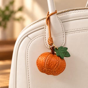 May include: A textured orange leather pumpkin charm with a green leaf accent featuring the initials A.G. engraved on it. The charm hangs from a tan leather strap attached to the handle of a smooth off-white handbag with visible stitching.