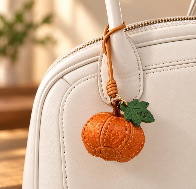 May include: A textured orange leather pumpkin charm with a green leaf accent featuring the initials A.G. engraved on it. The charm hangs from a tan leather strap attached to the handle of a smooth off-white handbag with visible stitching.