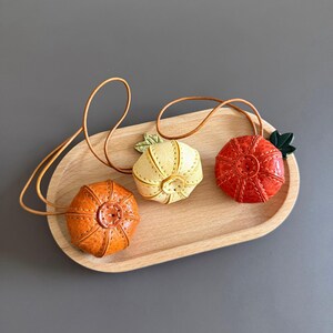 May include: Three small, round, pumpkin-shaped leather ornaments in orange, pale yellow, and red. Each piece features visible stitching, a central button detail, and a thin leather cord loop. A small green leaf is attached to the yellow and red items.