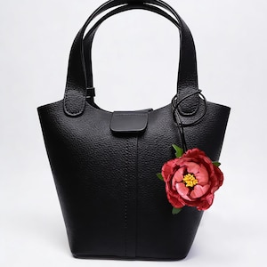 May include: Black leather tote bag with a textured finish, featuring two handles and a decorative red flower charm. The bag has a structured shape and a magnetic closure.