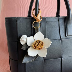 May include: A black leather tote bag with a woven design, featuring a white and brown flower-shaped keychain. The keychain has a gold-colored ring and a tan leather strap.