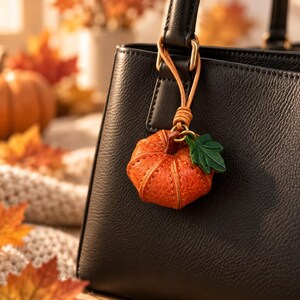 May include: A black leather handbag featuring a textured surface and a dangling pumpkin-shaped charm. The charm is crafted from orange leather with visible stitching, a brown stem, and a small green leather leaf attached to a thin leather cord.