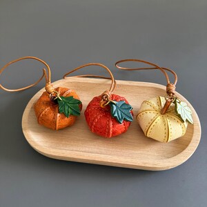May include: Three small pumpkin-shaped ornaments made of textured leather in orange, red, and cream colors. Each pumpkin features a green leather leaf charm attached to a thin brown leather cord loop for hanging, showcasing detailed stitching.