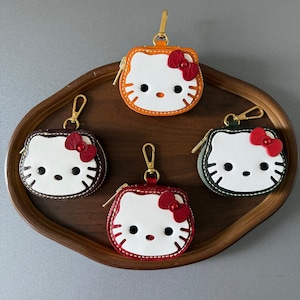 May include: A collection of five coin purses shaped like a cartoon cat face, each with a different colored border: orange, dark brown, green, red, and burgundy. Each purse has a red bow, black eyes, and a gold zipper and clasp. The purses are arranged on a wooden tray.