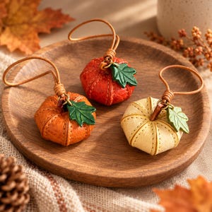 May include: Three small pumpkin-shaped ornaments crafted from textured leather in shades of orange, red, and cream. Each pumpkin features visible stitching, a small green leather leaf charm, and a thin leather cord loop for hanging purposes.