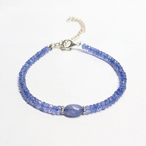 Può includere: Un braccialetto delicato con piccole perle di pietre preziose sfaccettate color blu pervinca disposte in cerchio. Una singola perla blu ovale, liscia e più grande è centrata, affiancata da due piccoli distanziatori in metallo argentato e una chiusura a moschettone.