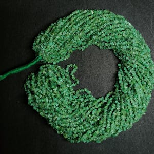 May include: A collection of small, irregular-shaped green emerald beads. The beads are strung together, forming a circular shape. The background is a solid black, providing contrast to the vibrant green gemstones.