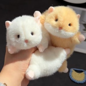 Puede incluir: Dos juguetes de peluche de hámster, uno blanco y otro naranja claro, se sostienen en una mano. Los juguetes tienen pequeñas orejas, narices y patas rosas. Los hámsters tienen ojos oscuros y pelaje suave y esponjoso. Un pequeño artículo de ganchillo azul y amarillo está en el fondo.