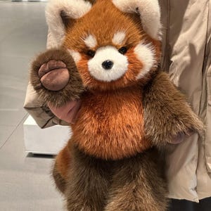 May include: A plush red panda stuffed animal with a fluffy, textured appearance. The toy features a mix of brown, orange, and cream-colored fur, with a detailed face and paws. The red panda is being held up, showcasing its soft, cuddly design.