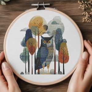 Puede incluir: Un bordado de punto de cruz con un búho azul en una escena de bosque con árboles en tonos azules, naranjas y amarillos. La obra de arte se muestra en un aro de madera redondo, mostrando un diseño artesanal con temática de la naturaleza.