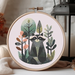 May include: A cross-stitch embroidery hoop featuring a raccoon in a forest scene. The design includes trees, foliage, and a raccoon with a black mask. The hoop is made of wood and is displayed with a lantern and pumpkin.