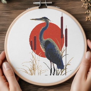 Könnte beinhalten: Ein Kreuzstich-Stickrahmen mit einem blauen Reiher vor einer roten Sonne, mit Rohrkolben und Schilf. Der Reiher ist mit Blau- und Grautönen detailliert. Die Stickerei befindet sich in einem Holzrahmen.