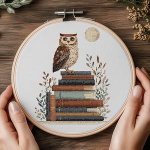 May include: A cross-stitch embroidery hoop featuring an owl perched atop a stack of books, with a full moon in the background. The owl has brown and beige feathers. The books are various shades of brown and blue. The embroidery is held in a wooden hoop.