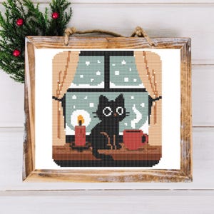 May include: A cross-stitch framed artwork depicts a black cat sitting on a windowsill, looking out at a snowy scene. A lit candle and a red mug sit beside the cat. The artwork is in a wooden frame, with a sprig of greenery in the corner.