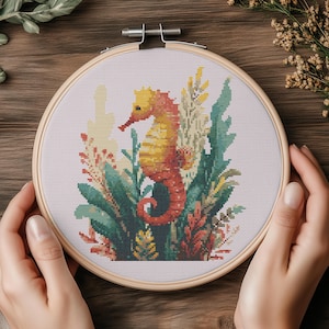 May include: Cross-stitch embroidery of a seahorse in a wooden hoop. The seahorse is orange and yellow, surrounded by green, yellow, and red plants. The embroidery is held by hands, with a wooden background.
