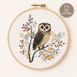 May include: A cross-stitch pattern featuring a brown and tan owl perched on a branch with light blue and red leaves, displayed in a round wooden embroidery hoop. A circular badge in the top right corner contains the text PDF PATTERN.
