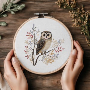 May include: Cross-stitch embroidery of an owl perched on a branch, set in a wooden hoop. The owl has brown and white feathers, with colorful leaves in the background. The embroidery is held by hands.