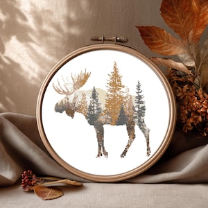 Puede incluir: Un bastidor de bordado de punto de cruz con la silueta de un alce y una escena de bosque. El diseño utiliza tonos de marrón, gris y dorado. El bordado está colocado en un bastidor de madera, creando una obra de arte rústica con temática de la naturaleza.